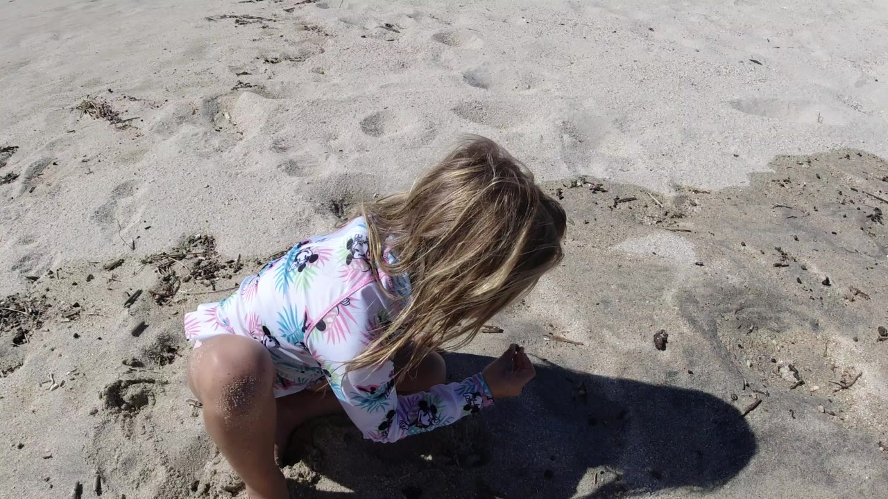 A young child kneels in pale sand in Los Cabos, Mexico, seen from above with hair covering the face and a dark shadow stretching across the frame.