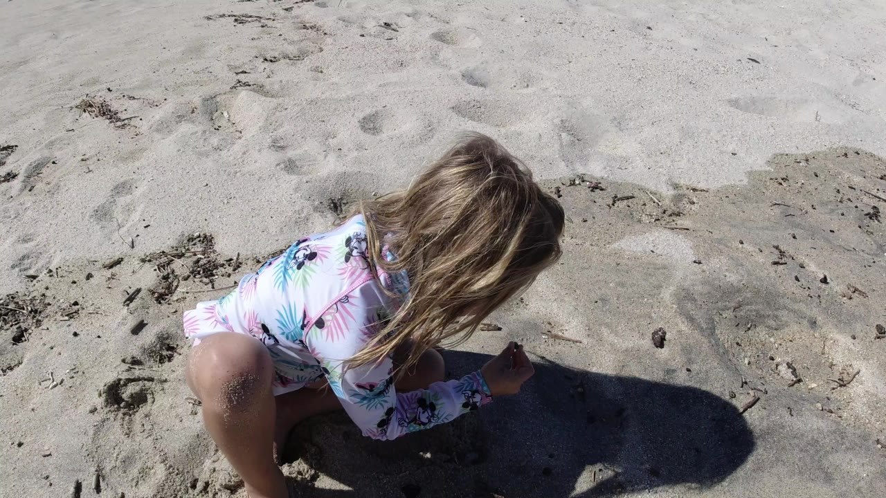 The child remains crouched over the Los Cabos sand, with the patterned swimsuit and windblown hair visible against footprints and small debris.