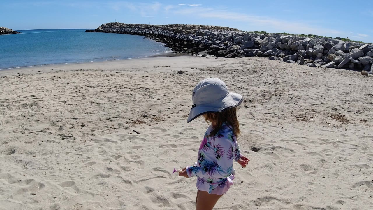 Young girl in a sun hat walking on a sandy beach near blue water and rocks.