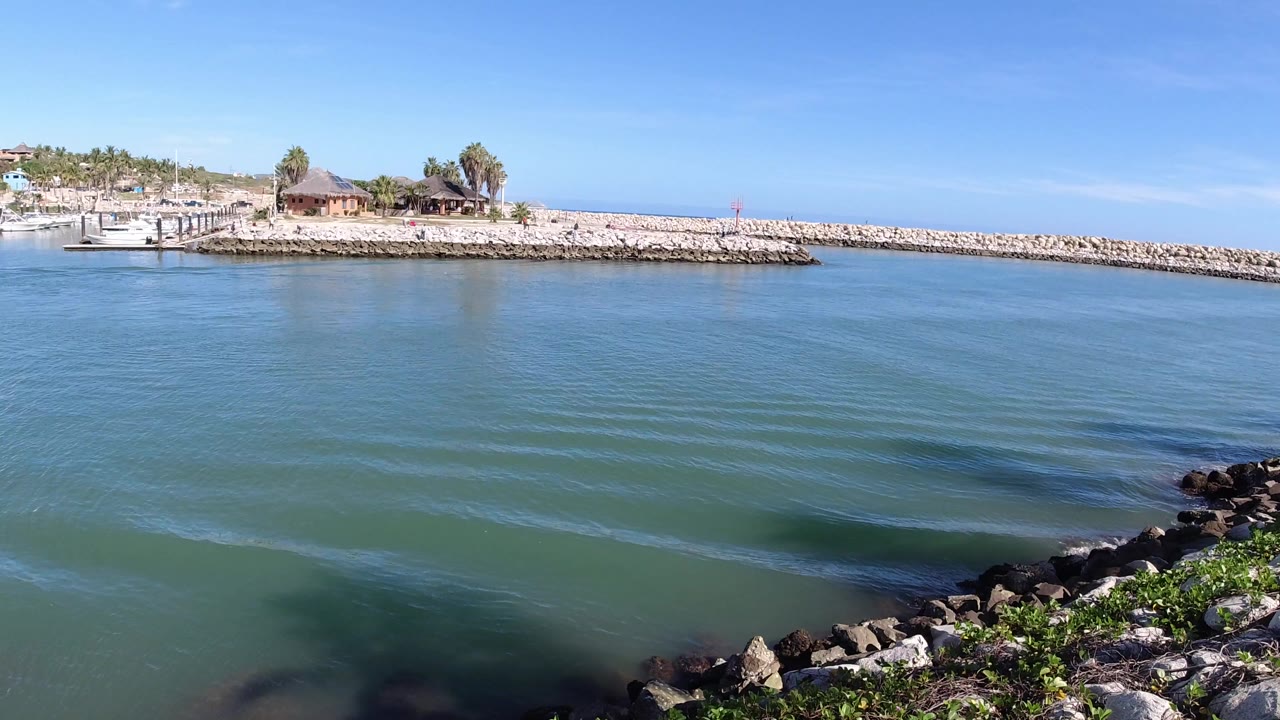 Opening frame of blue-green water in Los Cabos, Mexico, with shoreline rocks in the foreground, a stone breakwater and palapa-roof structures in the distance, and no visible people.