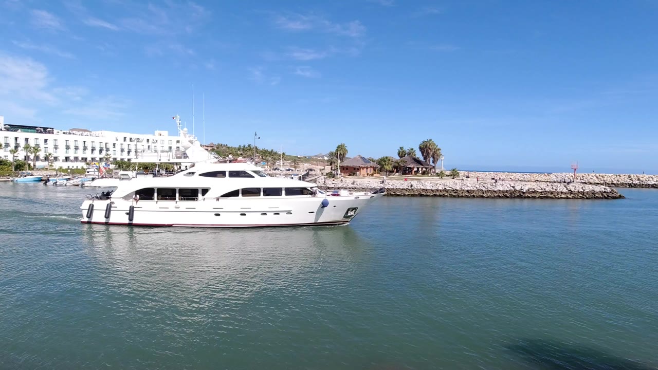 A white motor yacht sits left of center on blue-green water in Los Cabos, Mexico, with white shoreline buildings, palms, a rock breakwater, clear sky, and no visible people.