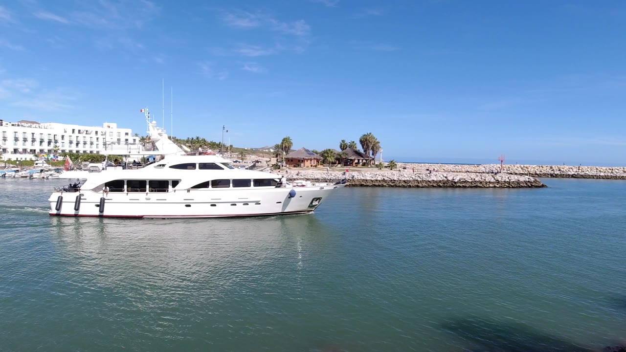 White yacht passing in front of a low rock breakwater under clear sky.