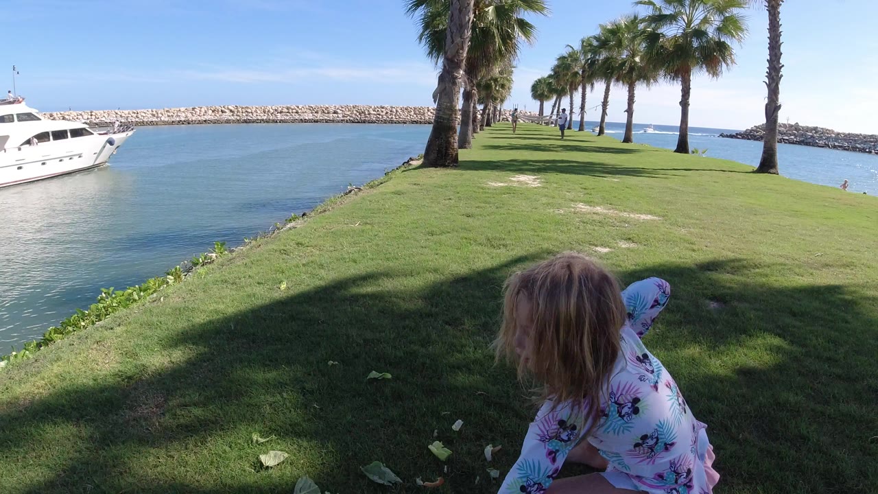 The girl remains low in the foreground grass while the white yacht sits on the left side of the marina channel and palm trees line the lawn in Los Cabos.