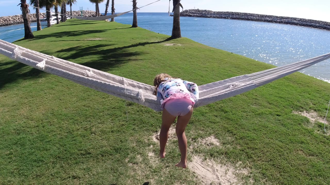 At 0:00 in Los Cabos, Mexico, a young girl bends toward a white hammock on bright grass with blue water, palm trees, and rock breakwaters behind her.