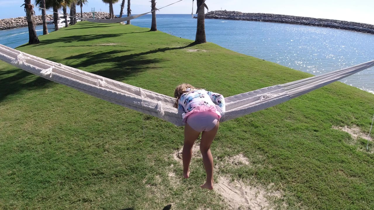 At 0:01 in Los Cabos, Mexico, the girl leans farther into the white hammock while standing on the grass near palm shadows and blue water.