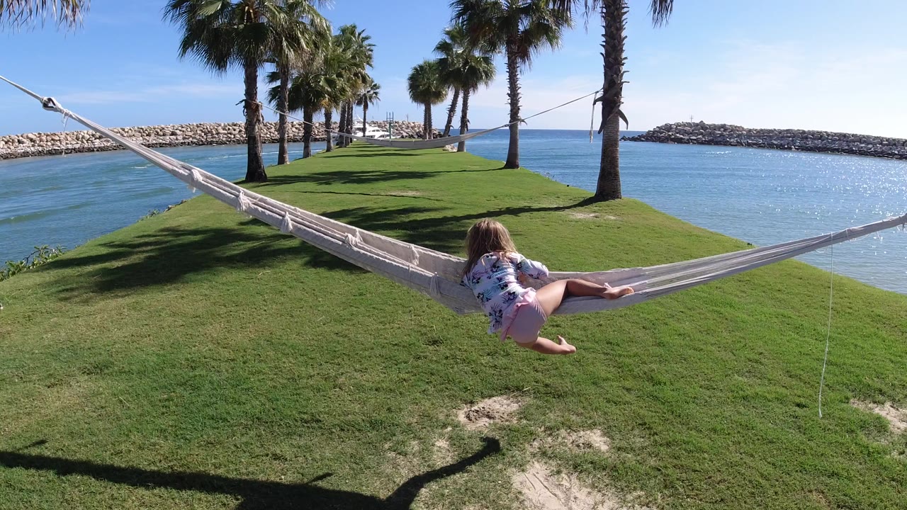 Child lying sideways in a hammock beside blue water and palm trees.