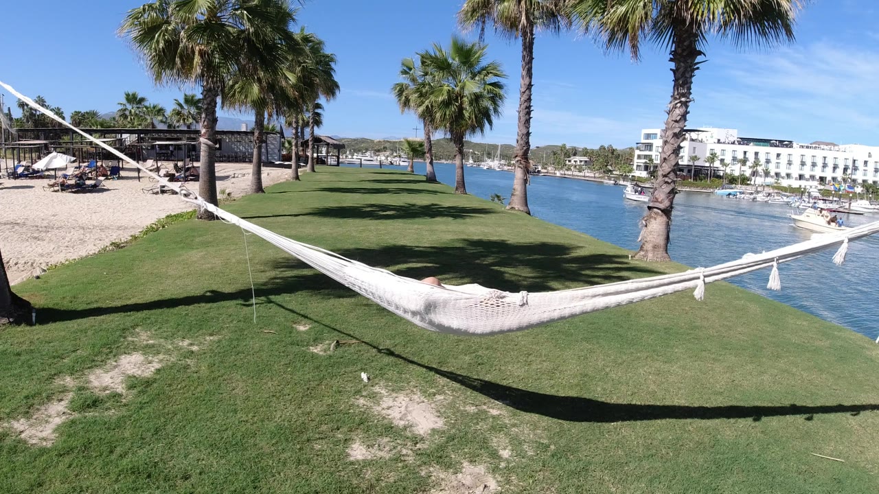 Opening frame in Los Cabos, Mexico, with a white hammock crossing a grassy edge between palms, sand and lounge chairs on the left, blue water on the right, and no visible people.