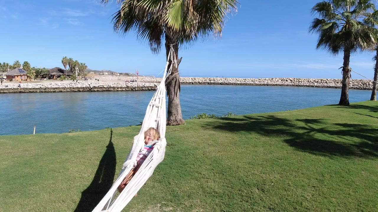 A young girl rests in a white hammock tied to a palm tree on a grassy lawn beside blue water in Los Cabos, Mexico, with a stone breakwater in the background.