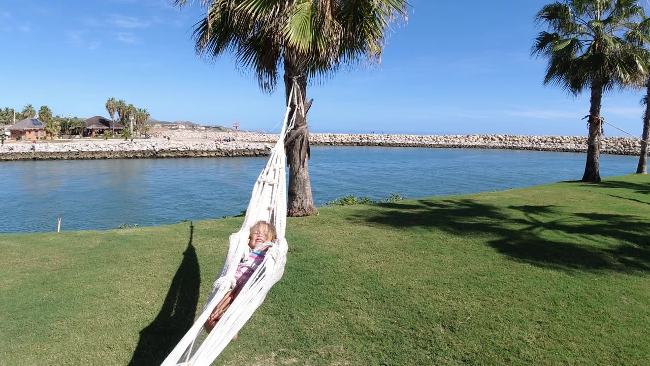 The girl remains visible in the white hammock as it hangs from the palm over grass, with calm blue water, a rock barrier, and low shoreline structures behind her in Los Cabos, Mexico.