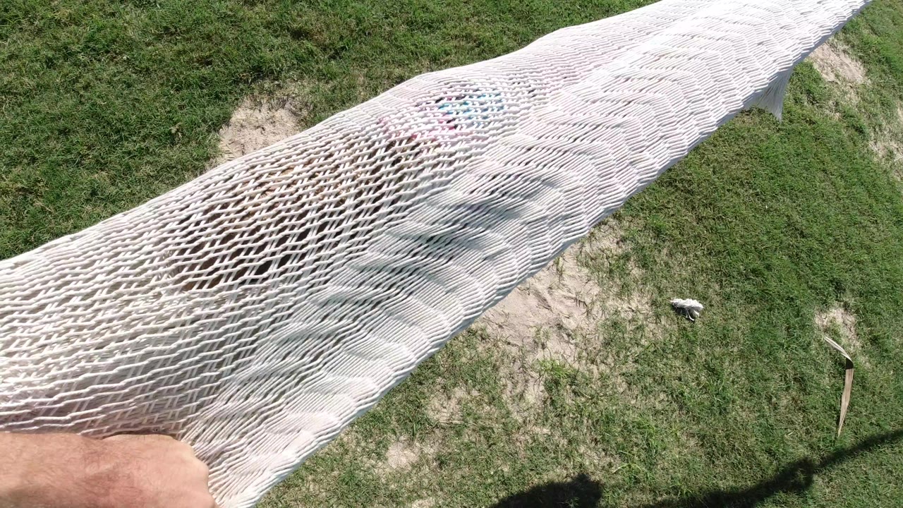 Overhead Los Cabos frame showing the white net swing crossing diagonally above grass, with an adult hand gripping the lower edge.