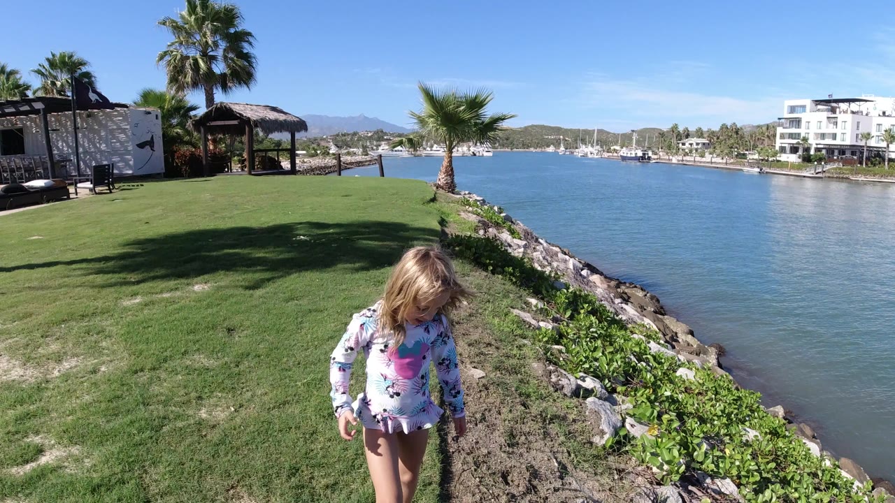 The girl continues along the grass and rocks beside the water in Los Cabos, Mexico, with a palm tree, small shaded structure, and distant boats across the channel.