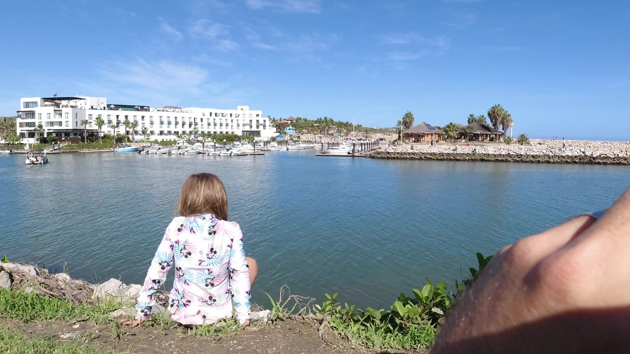 Young girl sitting beside blue water with a white hotel across the shore.