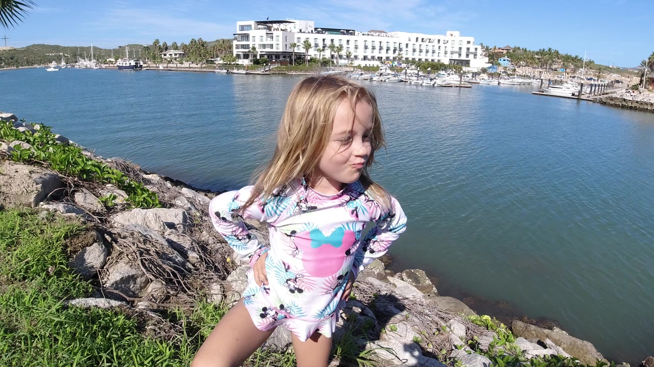 Blond child standing on rocks beside blue marina water.