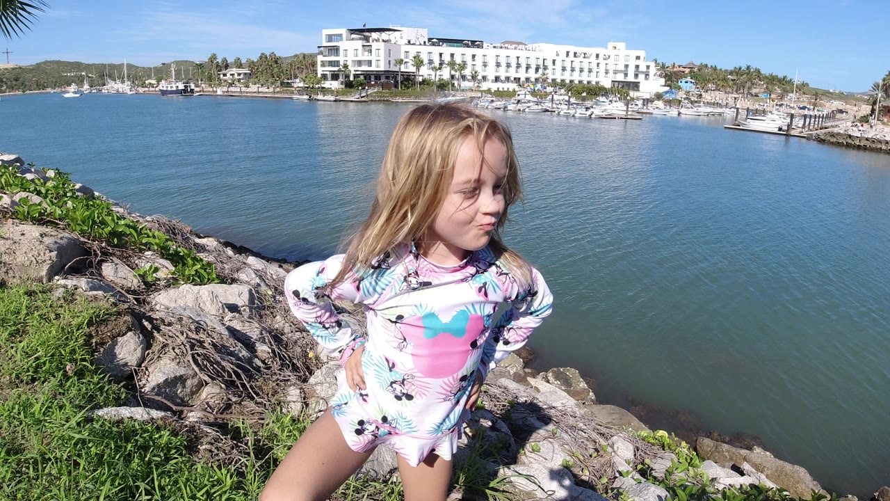 The girl remains posed on rocks beside the Los Cabos water, looking slightly to the side as the white marina-side building and moored boats fill the background.