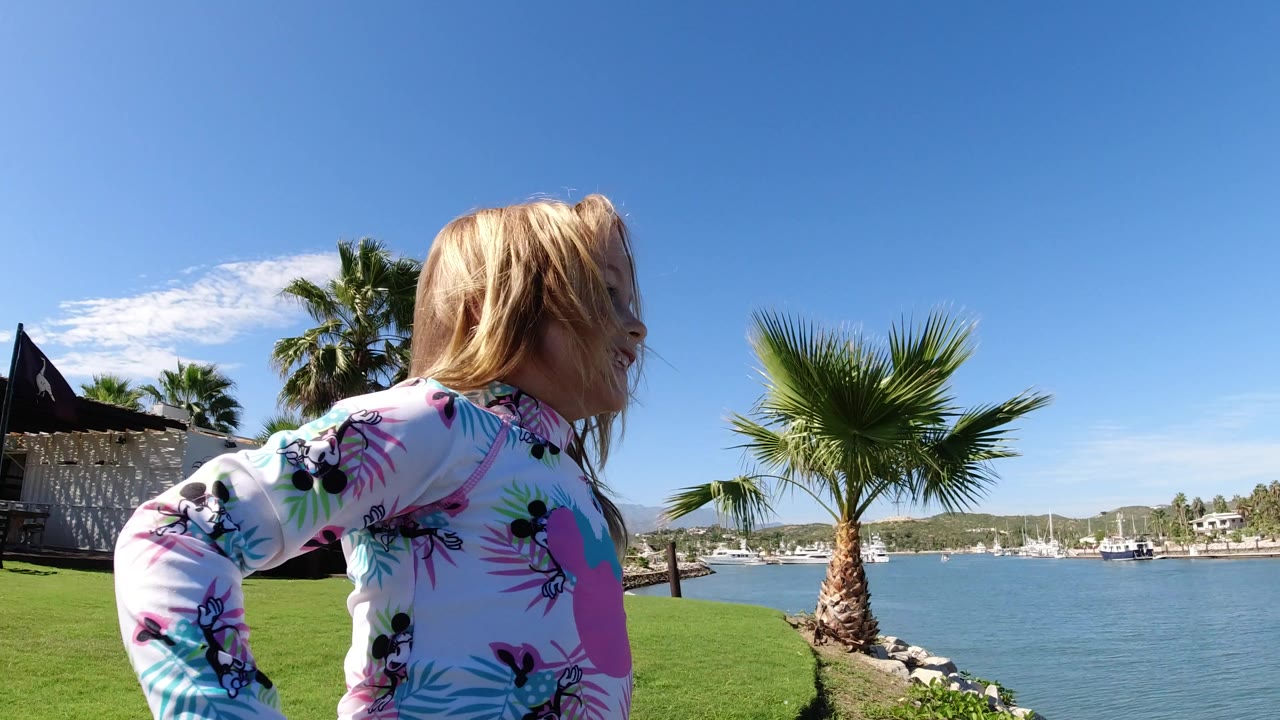 Blonde child in a colorful swim shirt near a waterfront lawn and palm trees.