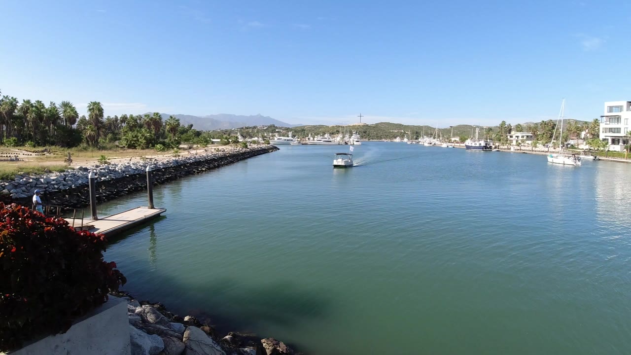 Canal view with a small boat, sailboats, rock breakwater, dock, and palms.
