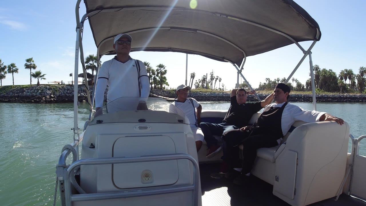 A covered boat in Los Cabos holds a standing man and seated passengers, with sunlight flaring across the canopy and calm water beside the hull.
