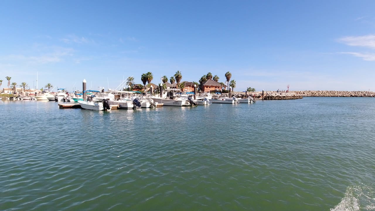 The second frame keeps the Los Cabos marina centered, with small boats along the dock, a rock breakwater in the distance, bright blue sky, and no visible people.