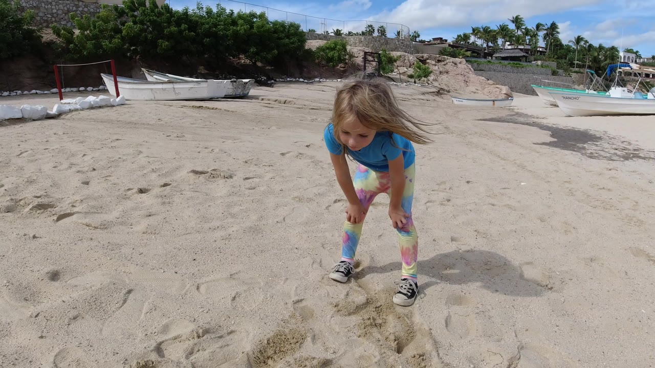 Young girl bending over sand on a beach with boats behind her.