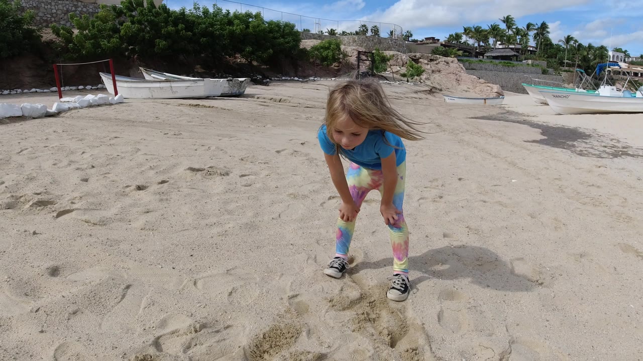 The girl remains bent forward over the sand in Los Cabos, framed low against a sandy foreground and background boats near trees.