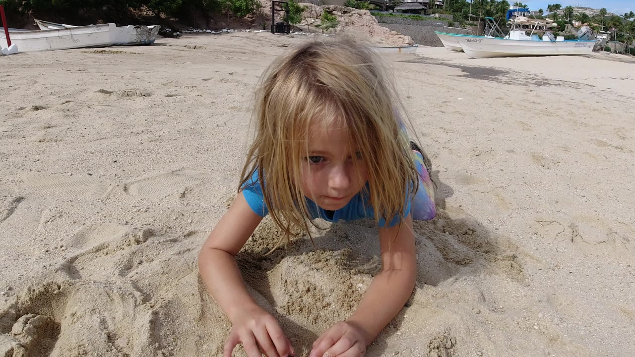 A blonde child in a blue shirt crouches close to the sand on a Los Cabos beach, with small boats and shrubs behind her.