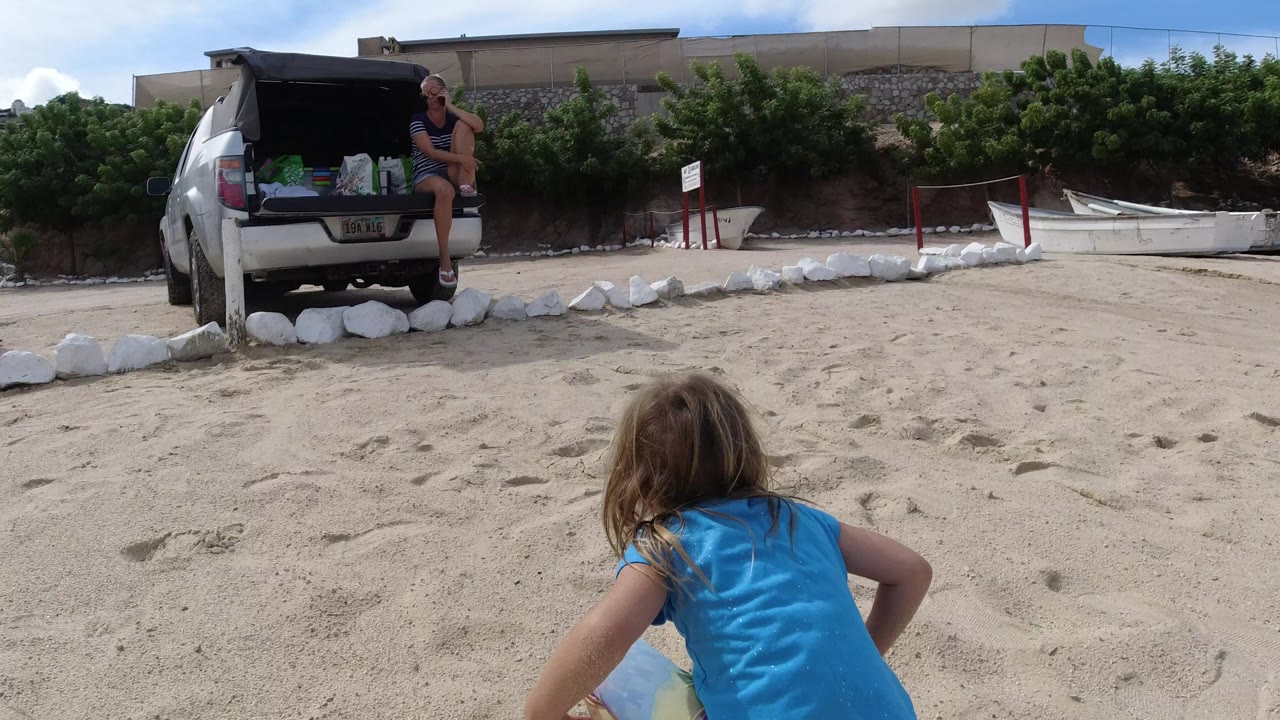 Little girl crouching in beach sand with a woman seated by a parked car behind her.