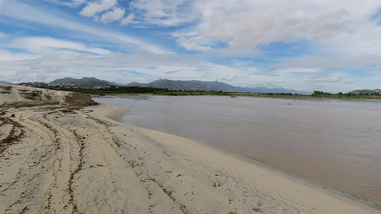 Curving sandbar beside shallow water with mountains and clouds beyond.