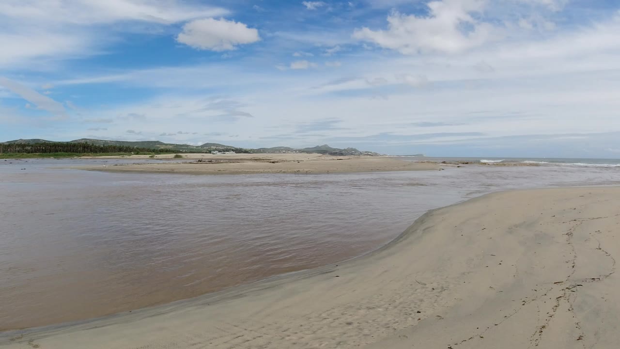 Los Cabos, Mexico low-tide beach at the start of the clip, with broad sand flats in the foreground, shallow water to the left, distant vegetation and hills, ocean on the horizon, and no visible people.