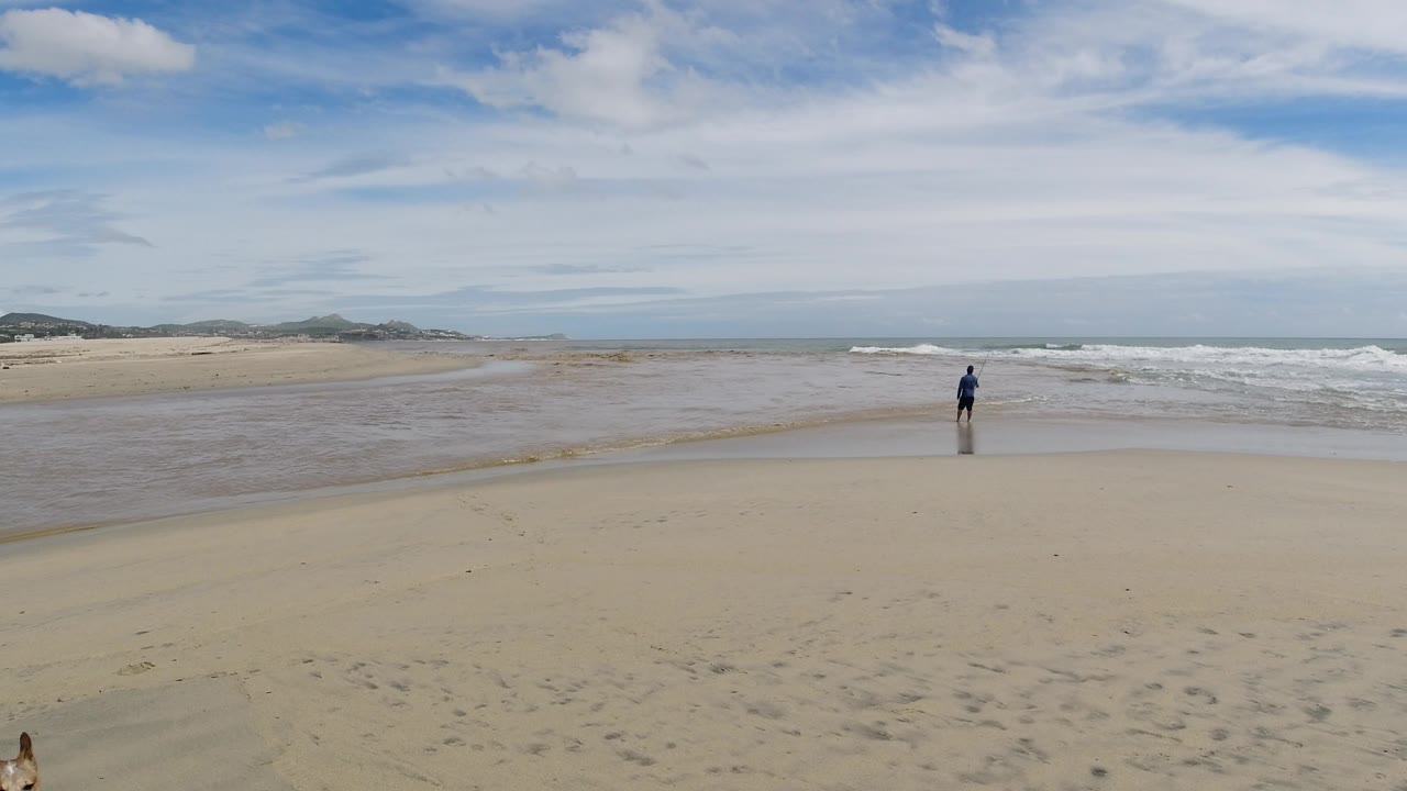 The man remains small in the frame near the incoming water, with a broad strip of wet sand and low surf stretching across the Los Cabos shoreline.
