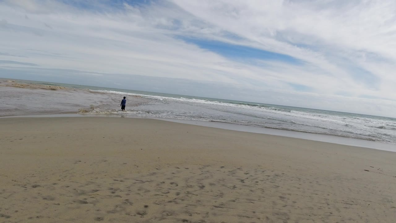 A distant man stands near the breaking surf on a wide sandy beach in Los Cabos, Mexico, with streaked clouds above and open sand in the foreground.