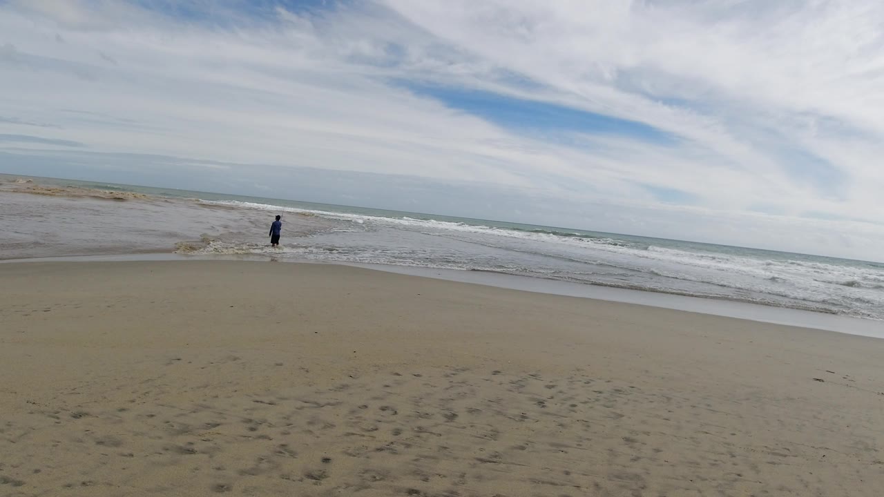 The lone person remains small near the waterline in Los Cabos, Mexico, as a band of foamy waves spreads across the shore beneath thin layered clouds.