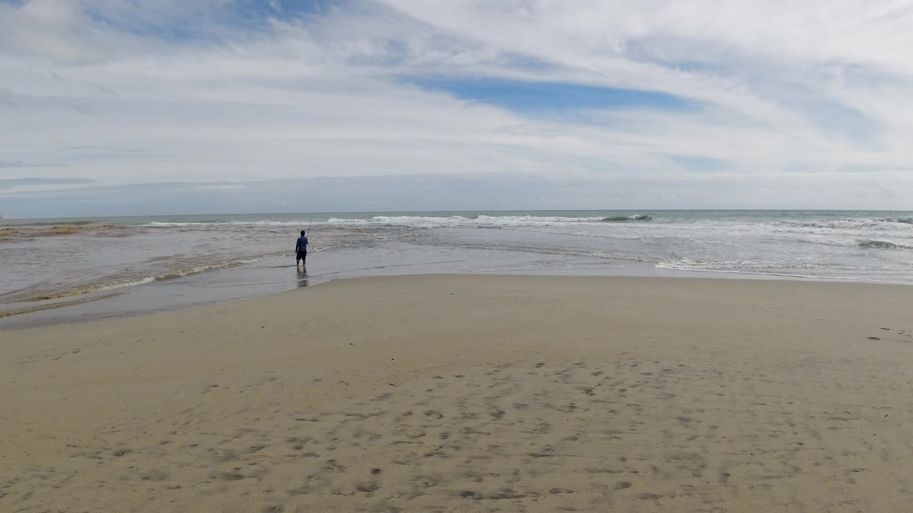 Distant person standing on a sandy beach near the surf under a cloudy sky.