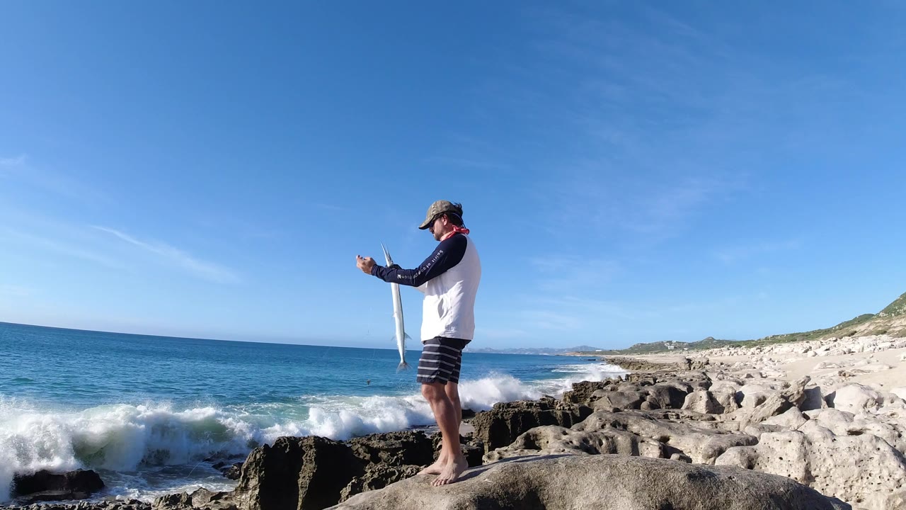 Man standing barefoot on a seaside rock holding a large fish near breaking waves.