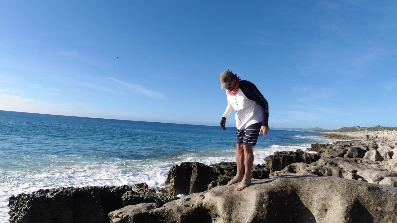 Barefoot man in a cap and long-sleeve shirt looks down while standing on pale shoreline rocks in Los Cabos, Mexico, with blue ocean and surf behind him.