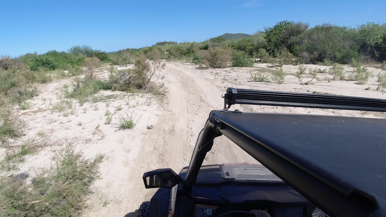 At the start of the Los Cabos clip, a sandy track runs ahead through low green scrub with the black Wrangler roof and light bar in the foreground and no visible people.