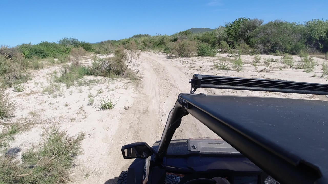 One second in, the Los Cabos track stays centered while the black roof edge and side mirror hold the lower foreground, with low brush and no visible people.