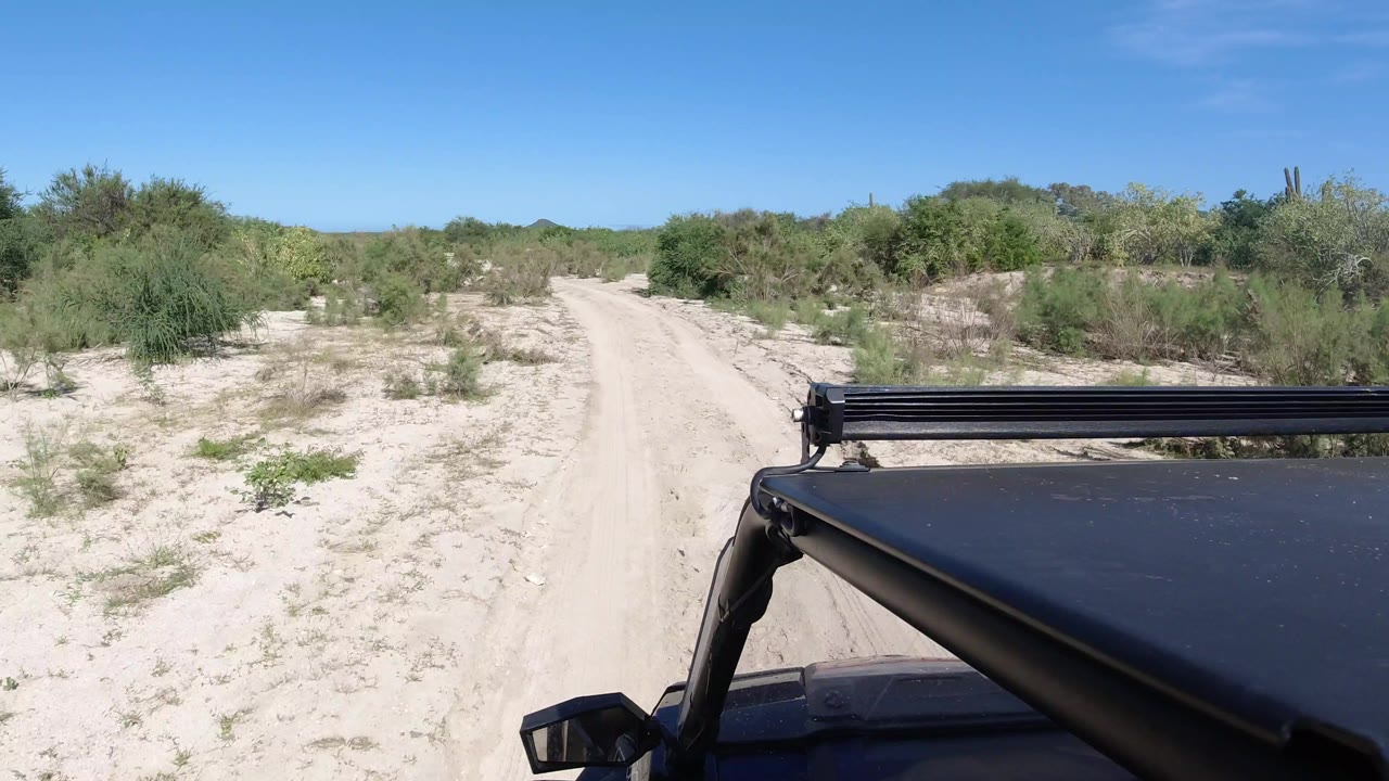 The vehicle continues along a narrow sandy road in Los Cabos, with scrub bushes on both sides, a clear blue sky, and no visible people.