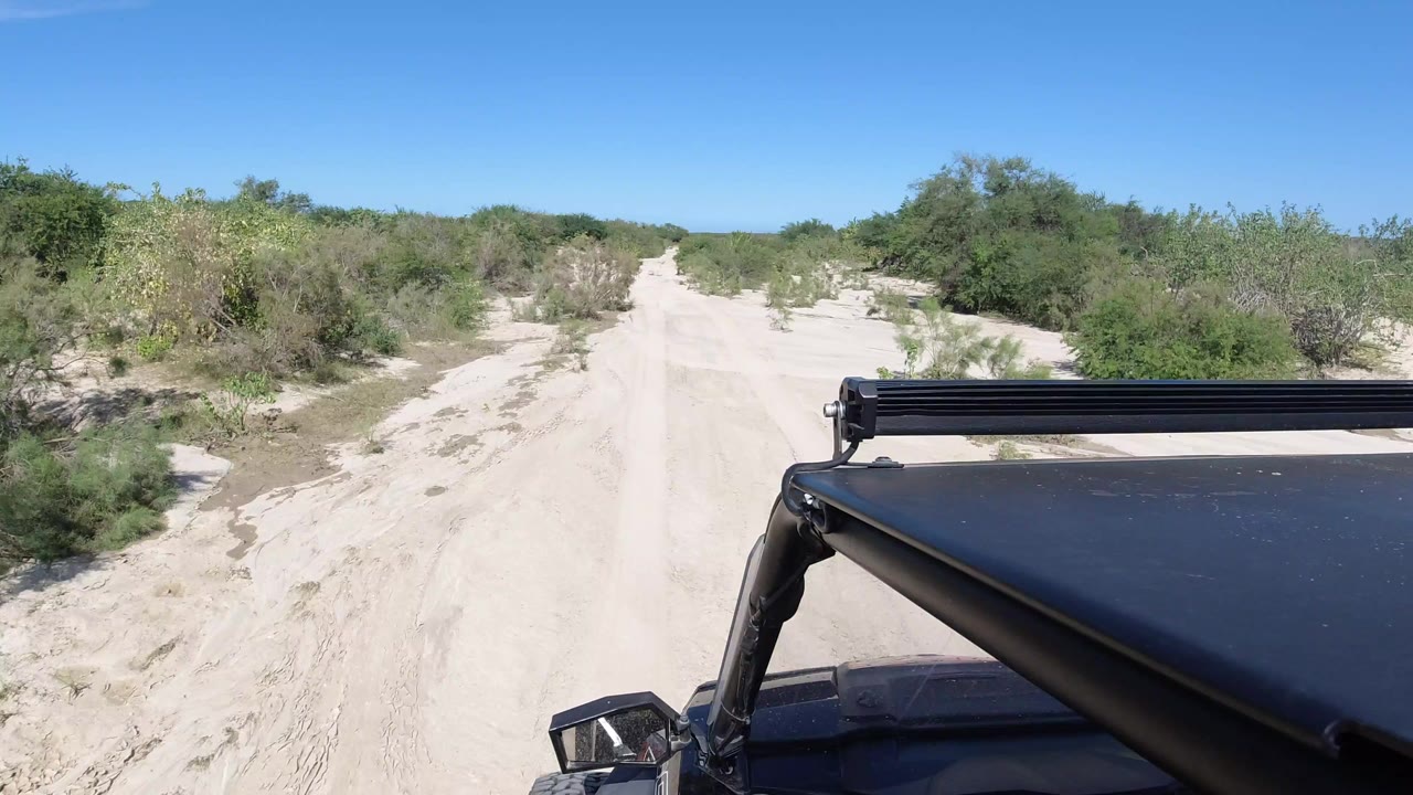 Forward vehicle view on a sandy road in Los Cabos, Mexico, with low green brush on both sides, clear blue sky above, black roof hardware in the foreground, and no visible people.
