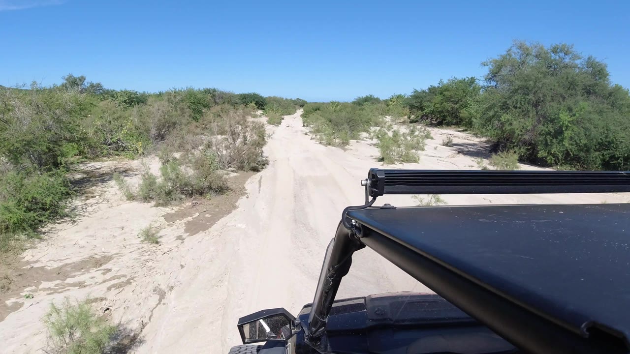 The sandy road narrows slightly through scrub in Los Cabos, Mexico, with brighter sand ruts ahead, dense brush at the sides, vehicle roof equipment at right, and no visible people.