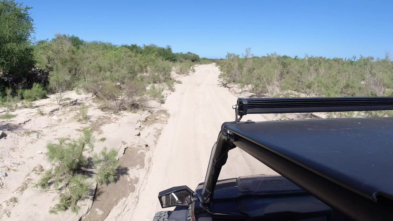 At 0:00 in Los Cabos, Mexico, the black Wrangler roof and light bar sit in the lower right as a pale sandy road opens ahead through low green scrub, with no visible people.