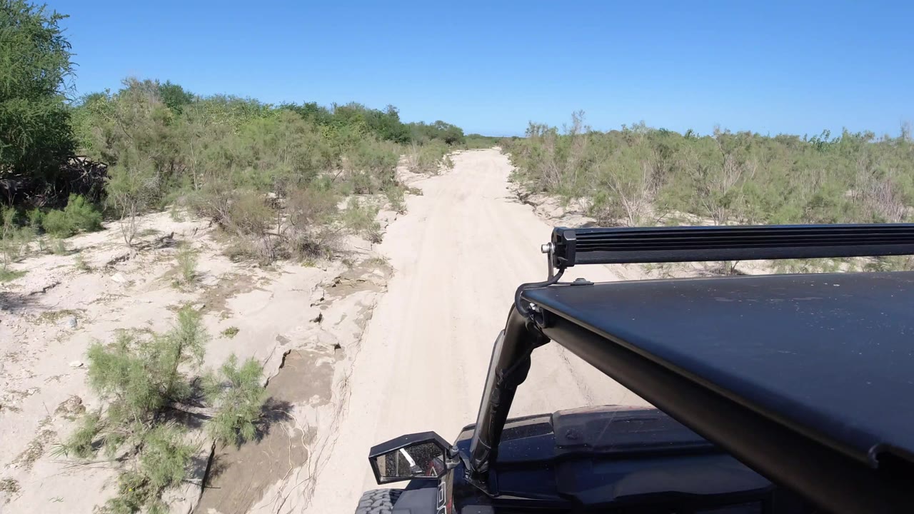 At 0:01 in Los Cabos, Mexico, the road appears slightly closer and straighter ahead, bordered by sandy banks and brush, with the black Wrangler still anchored in the lower right and no visible people.