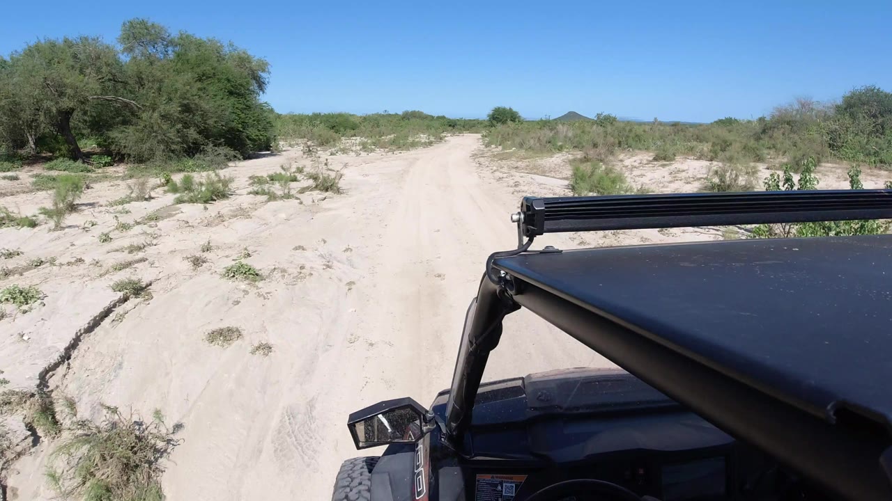 One second in, the vehicle foreground remains on the right as the sandy Los Cabos road continues forward between scrubby plants and a clear blue sky, no visible people.