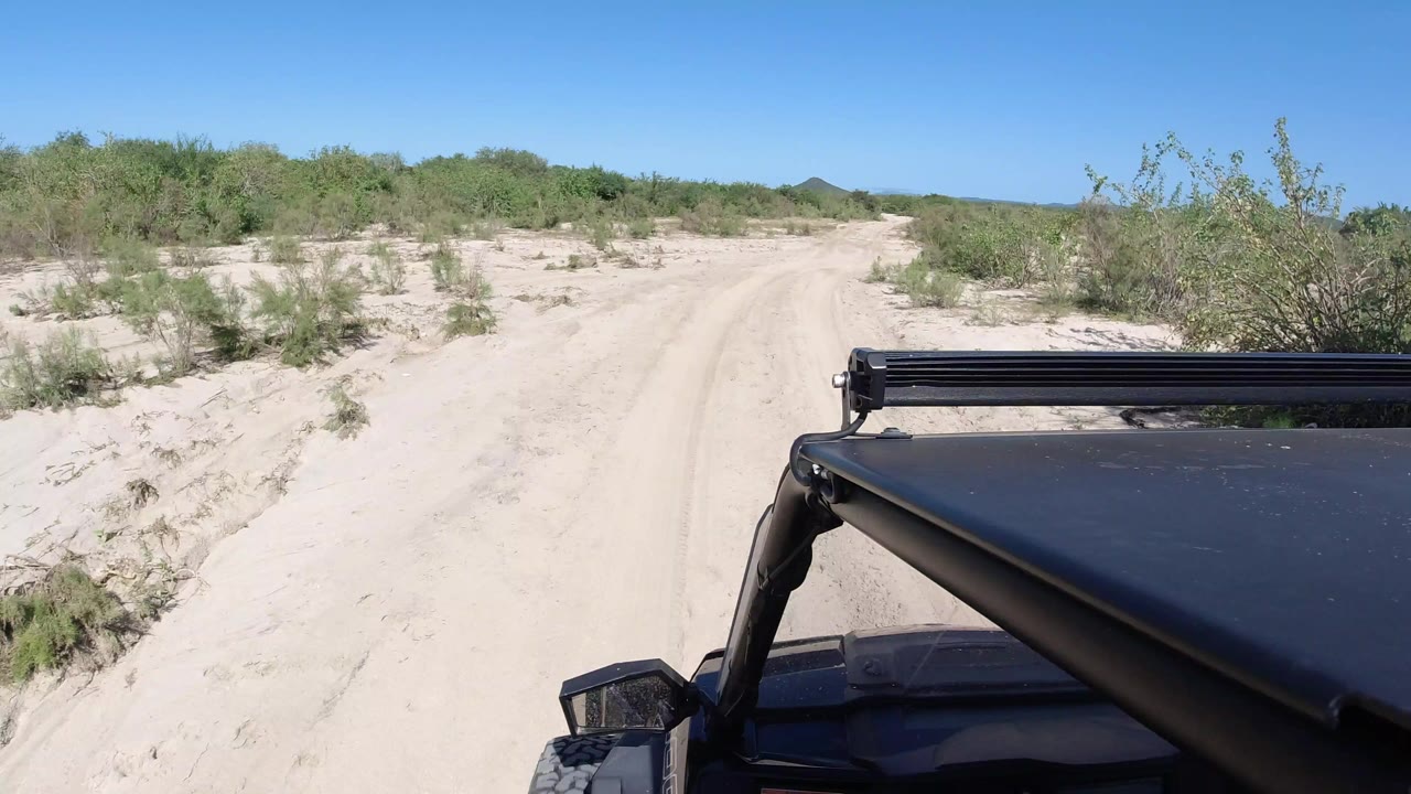 Start of a Los Cabos road view with a black Jeep hood and light bar in the foreground, pale sandy track ahead, low green brush, distant hills, and no visible people.
