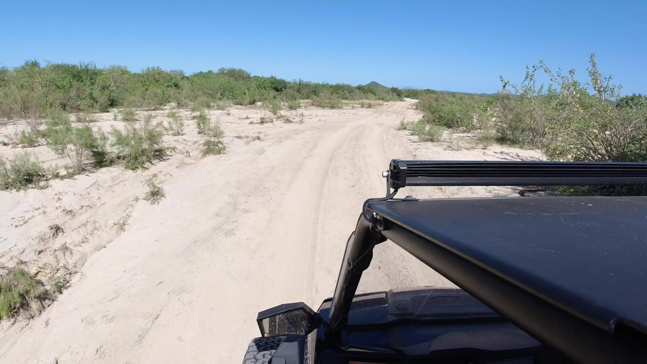 A forward Los Cabos driving view shows the Jeep closer to the right edge as the sandy road continues between scrub plants under clear sky, with no visible people.