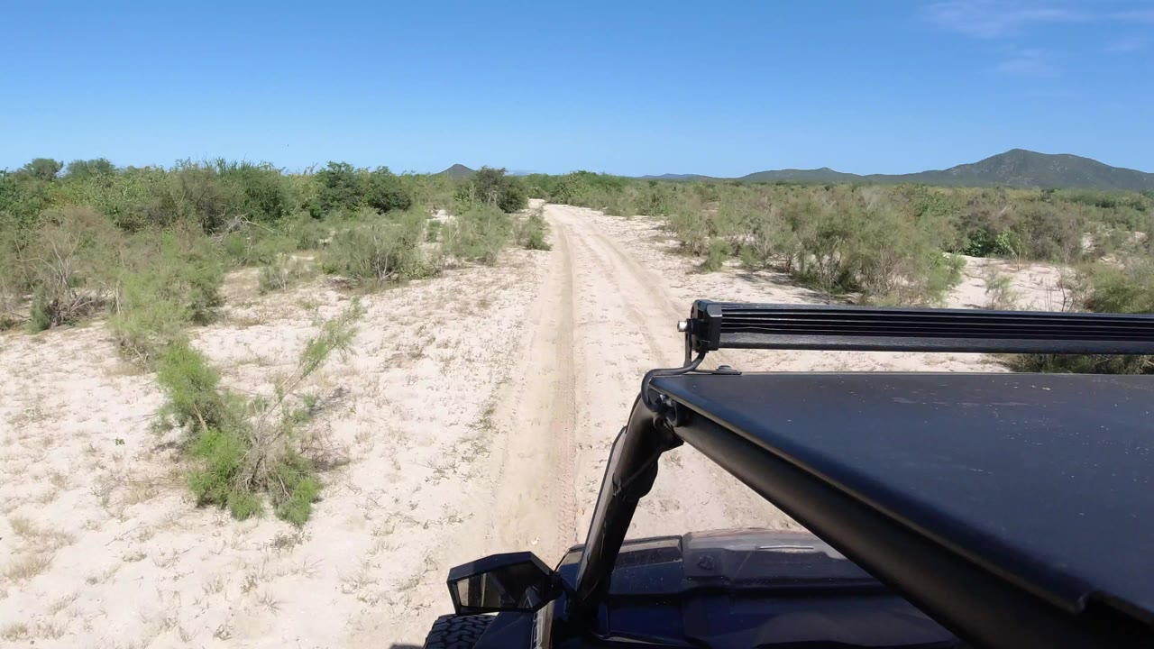 Forward view from a vehicle on a pale sandy road in Los Cabos, Mexico, with low green brush, distant hills, blue sky, and no visible people.