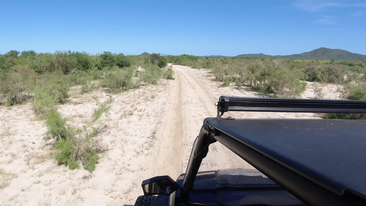 A similar forward Los Cabos road view shows the black vehicle roof at lower right, the track ahead, scattered scrub, low mountains, and no visible people.
