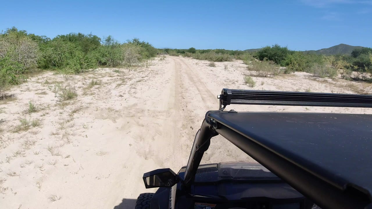 Opening view of a sandy single-track road in Los Cabos, Mexico, bordered by green brush with low hills ahead and no visible people.