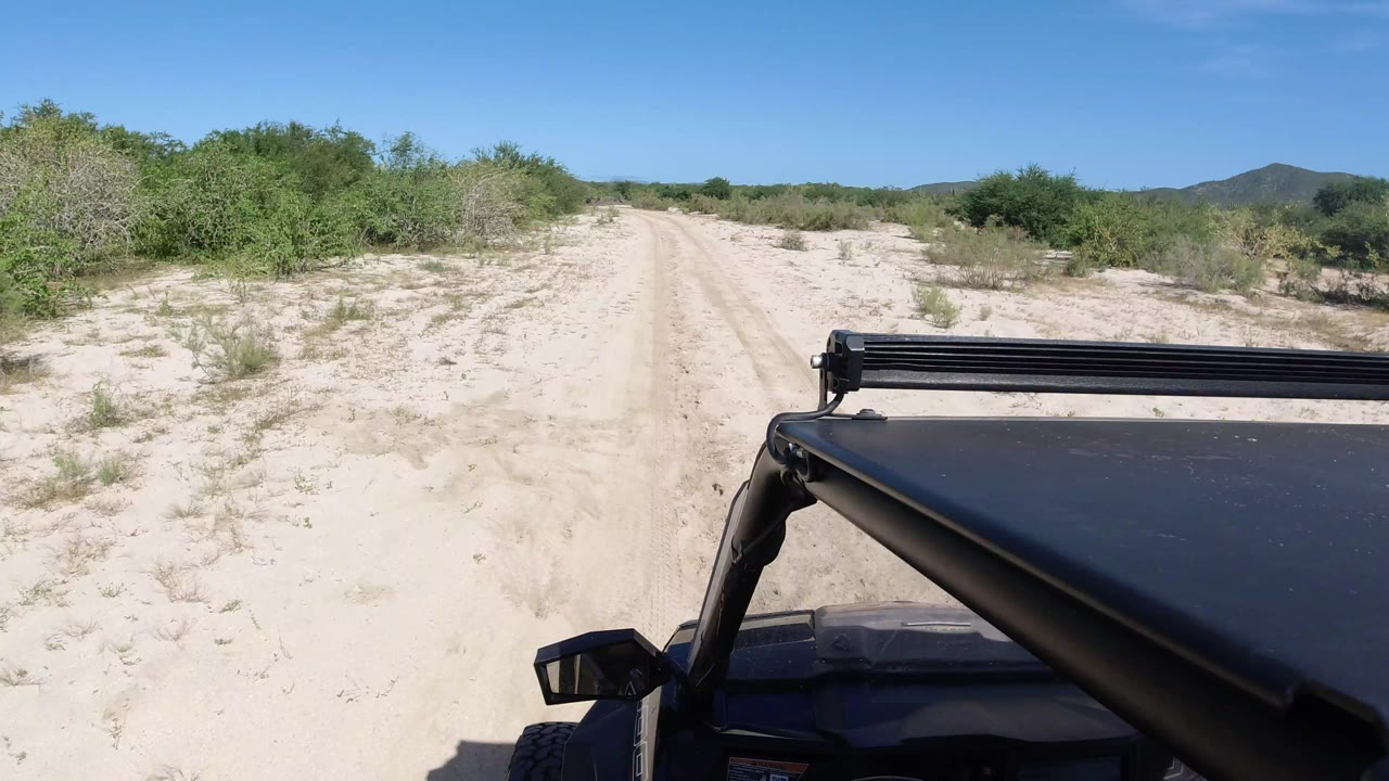 A pale road continues through low green vegetation in Los Cabos, Mexico, with the black vehicle roofline and light bar visible at lower right and no visible people.