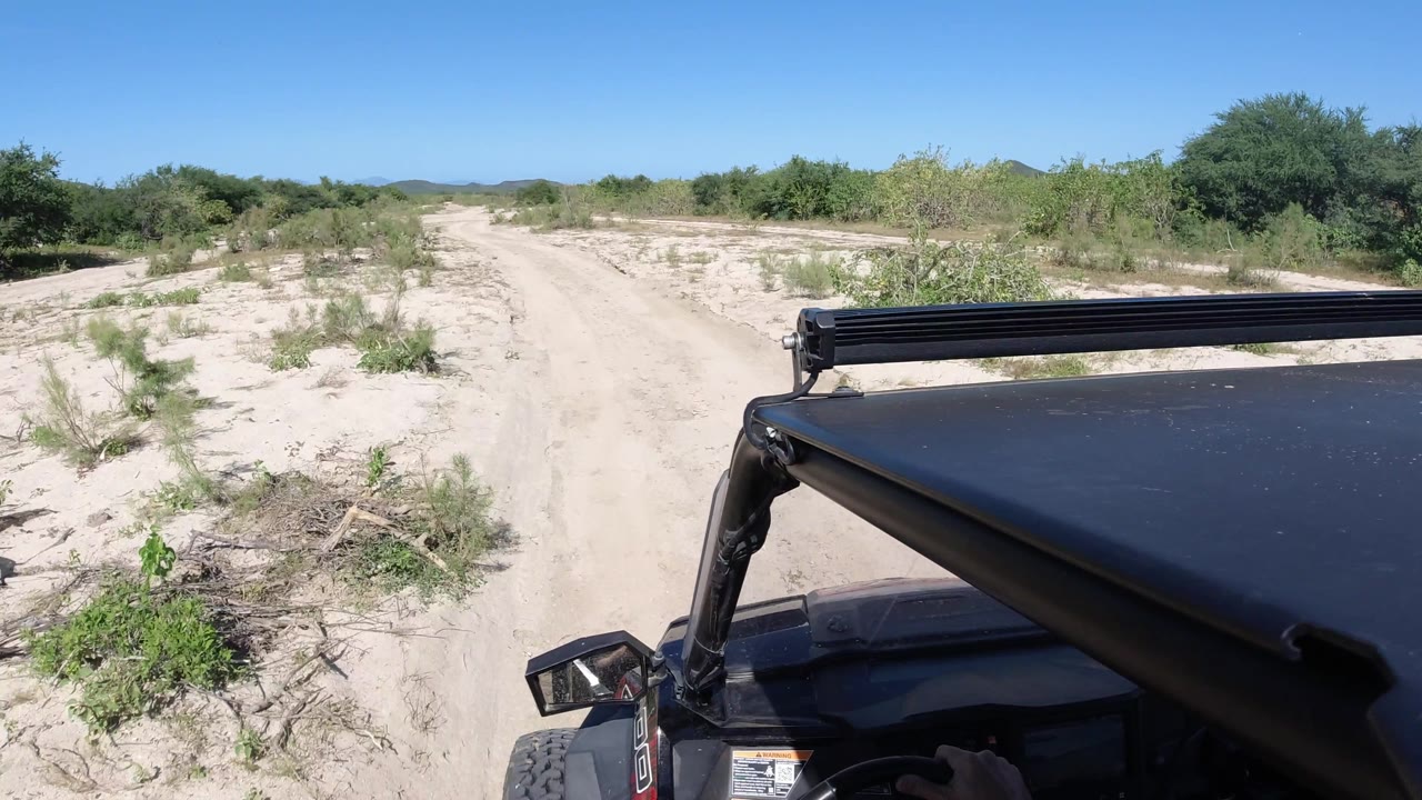 Dusty forked dirt path with shrubs and vehicle parts at the lower right.