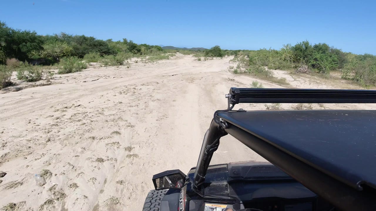 Vehicle POV in Los Cabos looking across pale sand tracks, scattered brush, a low horizon, and the black roof and light bar at lower right with no visible people.
